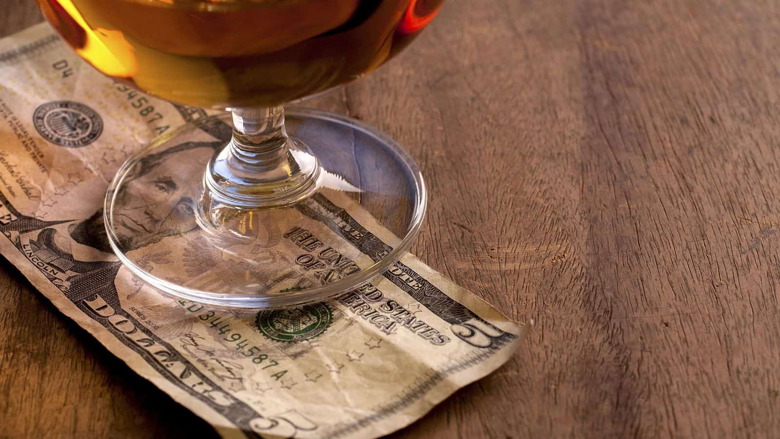 A glass with a drink is placed on a five-dollar bill on a wooden table. The drink appears to be an amber liquid, possibly whiskey or brandy. The dollar bill is partially visible beneath the glass.