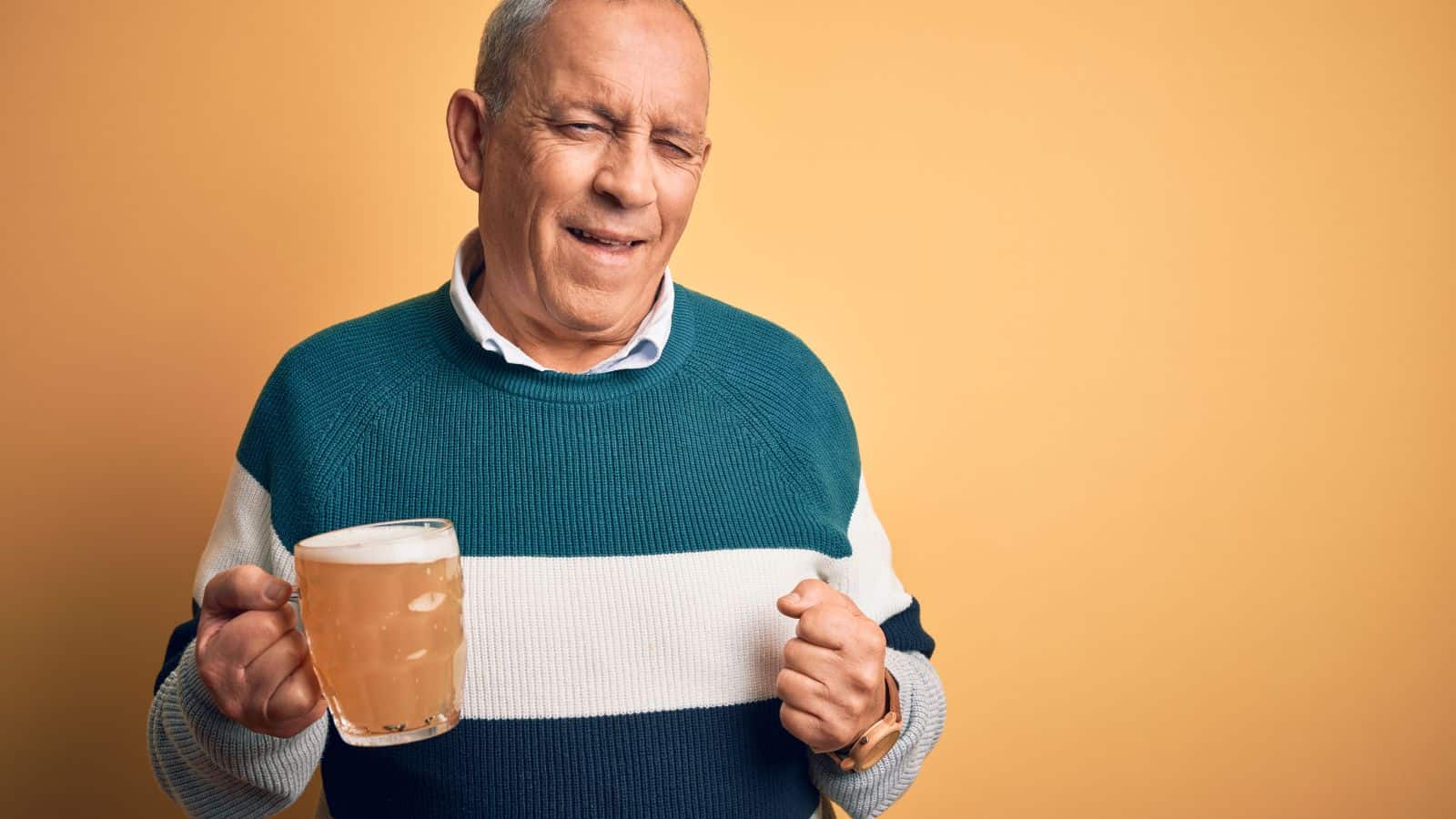 An older man with short hair holds a mug of beer and winks at the camera. He is wearing a teal, white, and gray striped sweater. The background is a solid yellow color.