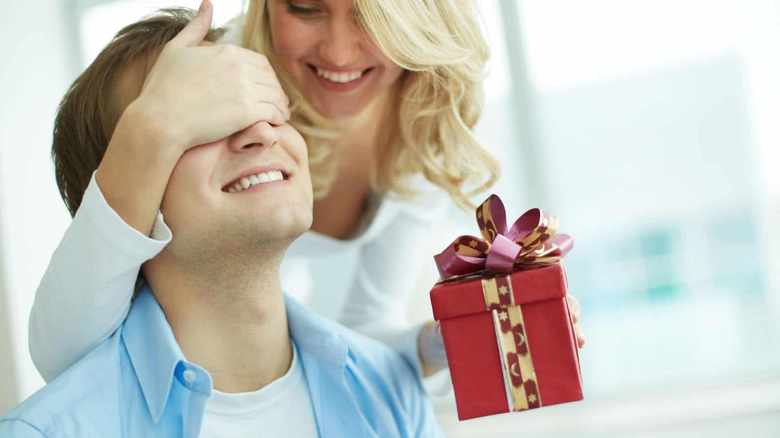 A smiling man is sitting while a woman stands behind him, covering his eyes with one hand. She holds a small red gift box with a ribbon in the other hand. Both appear happy, and the setting is brightly lit.