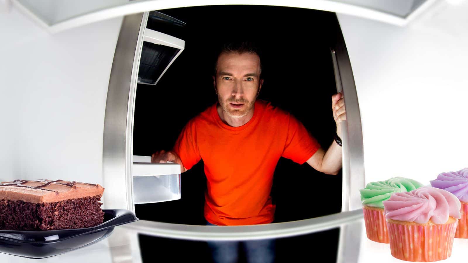 A person wearing a red shirt is looking into a refrigerator. Inside, there is a piece of chocolate cake on the left and colorful cupcakes on the right. The interior is dimly lit, highlighting the desserts.