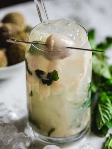 A tall glass filled with a light-colored cocktail containing lychee and mint leaves, garnished with a lychee on a pick. Ice cubes surround the glass. A bowl of lychees and fresh mint leaves are in the background on a marble surface.
