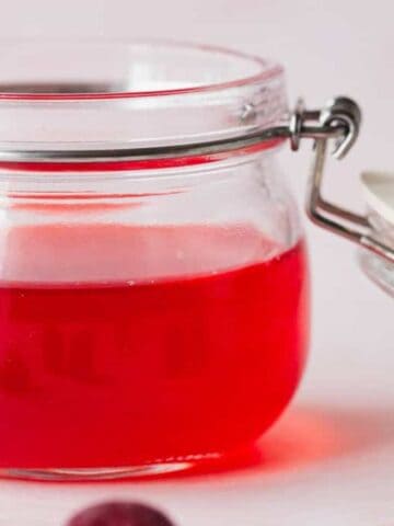 A glass jar filled with red jelly is shown with the lid resting open beside it. The jar is placed on a light surface, and a few cranberries are scattered around.