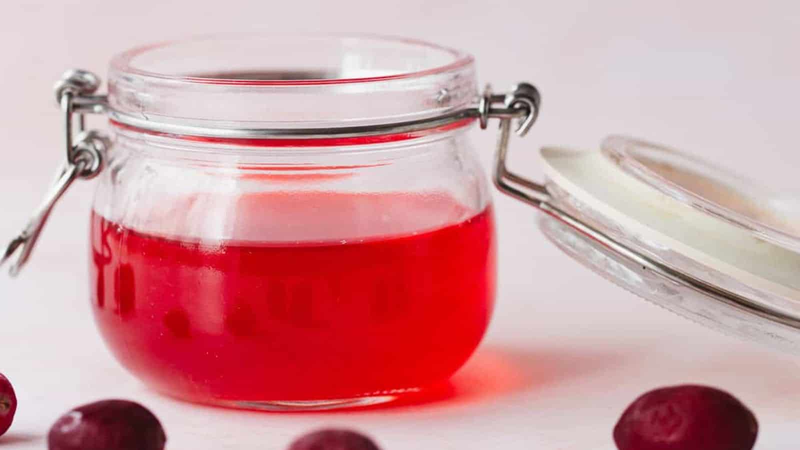 A glass jar filled with red jelly is shown with the lid resting open beside it. The jar is placed on a light surface, and a few cranberries are scattered around.