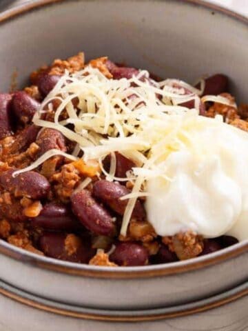 A bowl of chili with ground meat, kidney beans, and tomato sauce, topped with shredded cheese and a dollop of sour cream. The bowl is stacked with another on a plate, and two metal spoons are placed beside it.