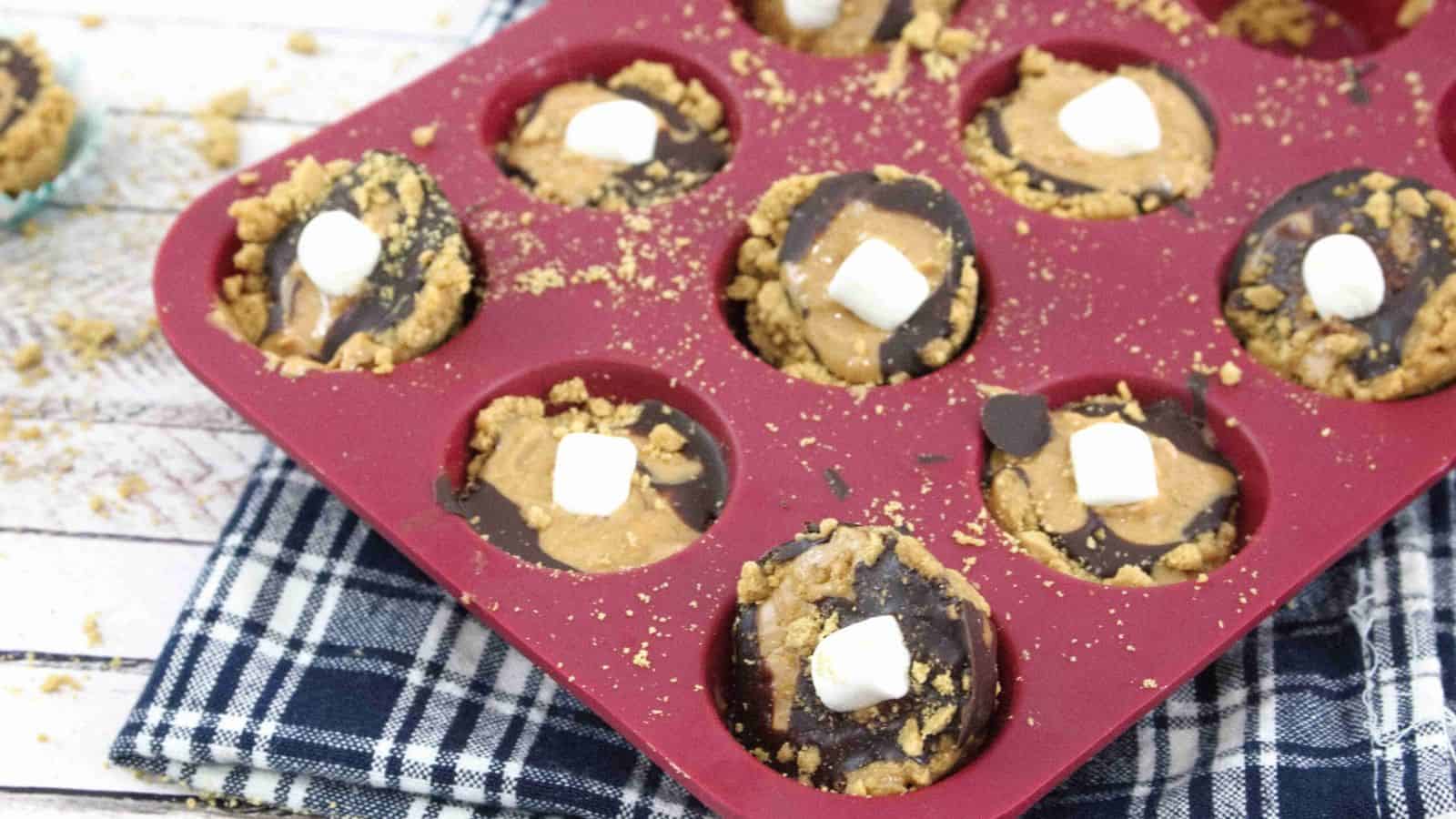 Mini s'mores cups with graham cracker crust, chocolate, peanut butter and mini marshmallows in a mini, silicone red muffin tin on a blue linen.