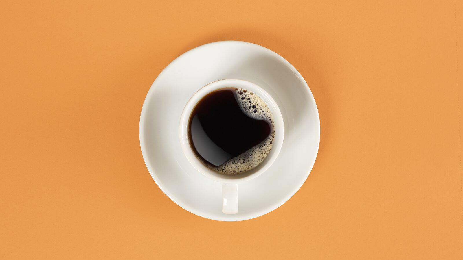 A white cup filled with black coffee sits on a matching saucer against an orange background. The coffee has a light layer of bubbles on one side. The image is shot from above.