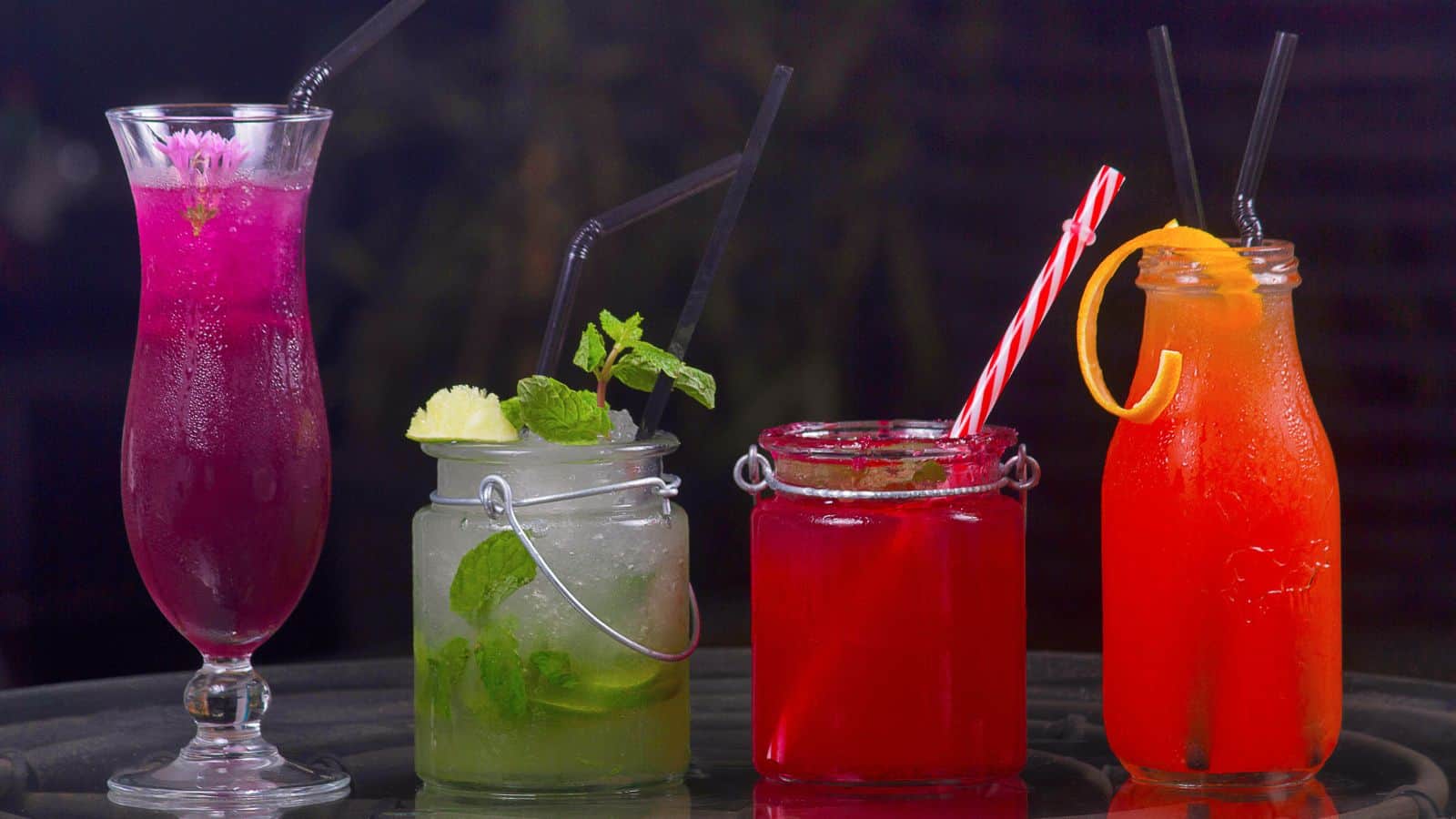 Four colorful drinks on a table. From left to right: a purple drink in a tall glass with a flower, a green drink with lime and mint in a jar, a red drink with a candy cane straw in a jar, and an orange drink with an orange slice in a glass.