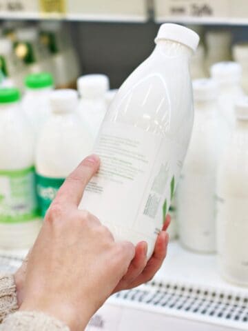 A person holds a white plastic bottle while shopping in a dairy aisle. Several similar bottles with green and white caps are displayed on the shelves in the background. The person appears to be reading the label of the bottle.