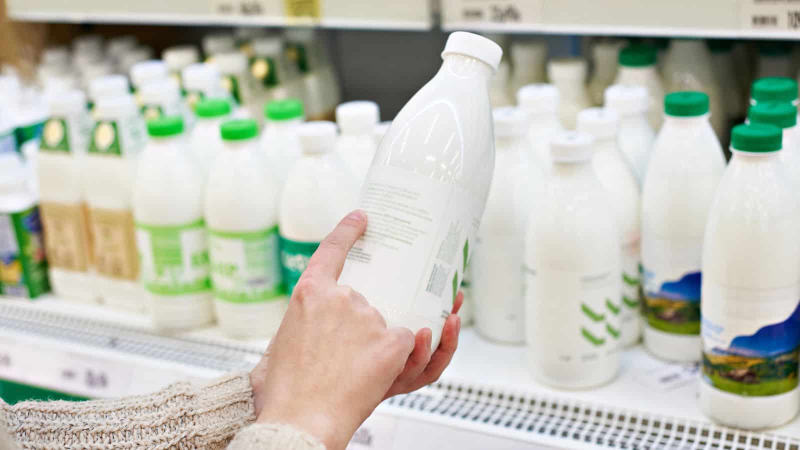 A person holds a white plastic bottle while shopping in a dairy aisle. Several similar bottles with green and white caps are displayed on the shelves in the background. The person appears to be reading the label of the bottle.