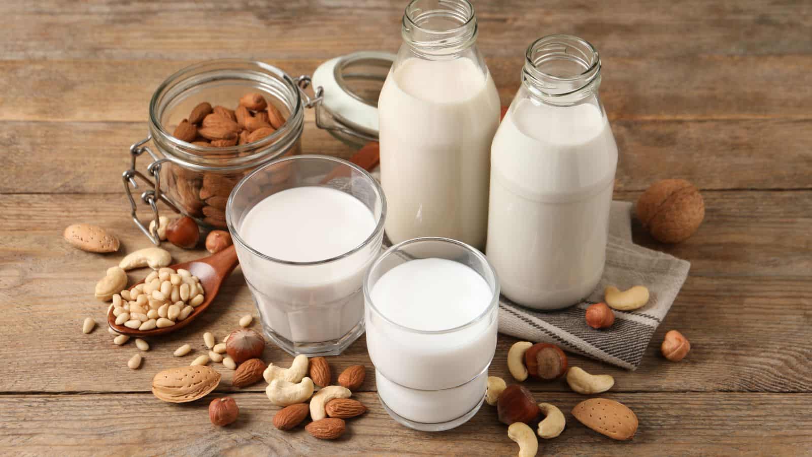 Various glass bottles and glasses of plant-based milk are displayed on a wooden surface. A jar of almonds, a spoon with soybeans, and assorted nuts like cashews and hazelnuts are scattered around, emphasizing the ingredients.