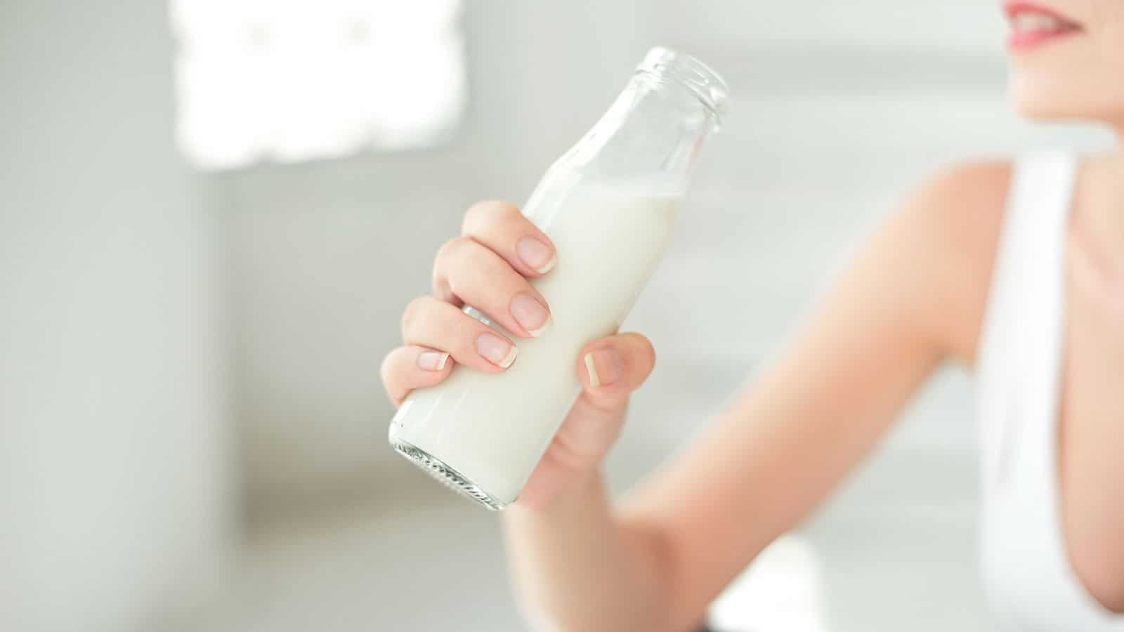 A person holds a glass bottle filled with milk in their hand. The background is bright and blurred, and the person is wearing a white sleeveless top. The focus is on the bottle and the person's hand.