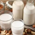 Two glass bottles and two glasses of milk are on a wooden surface. Various nuts, including almonds, cashews, and hazelnuts, are scattered around. A glass jar with a few nuts inside is open in the background.