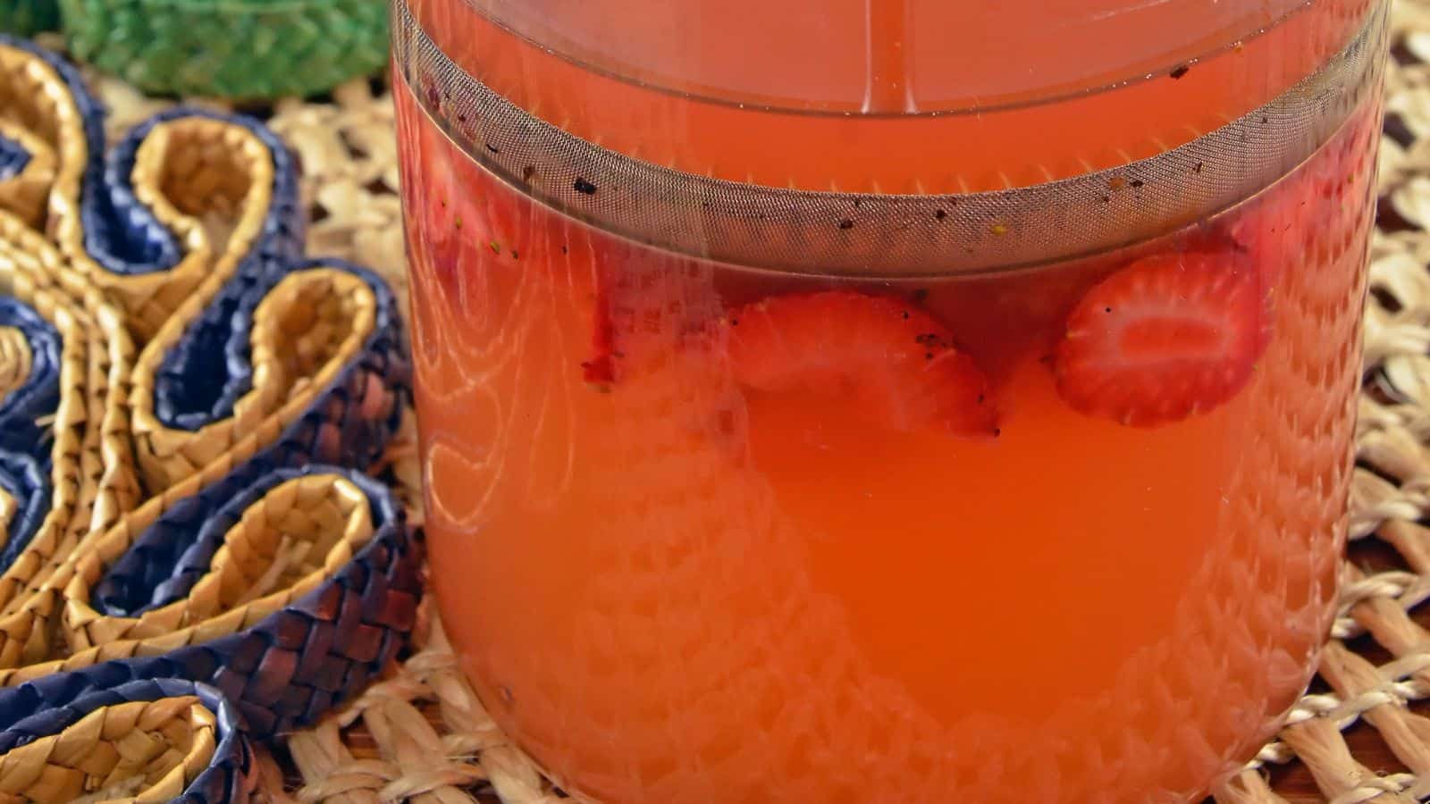 A glass container filled with pink lemonade and sliced strawberries. The container has a metal spout. It sits on a woven surface next to a textured, multicolored decorative object.