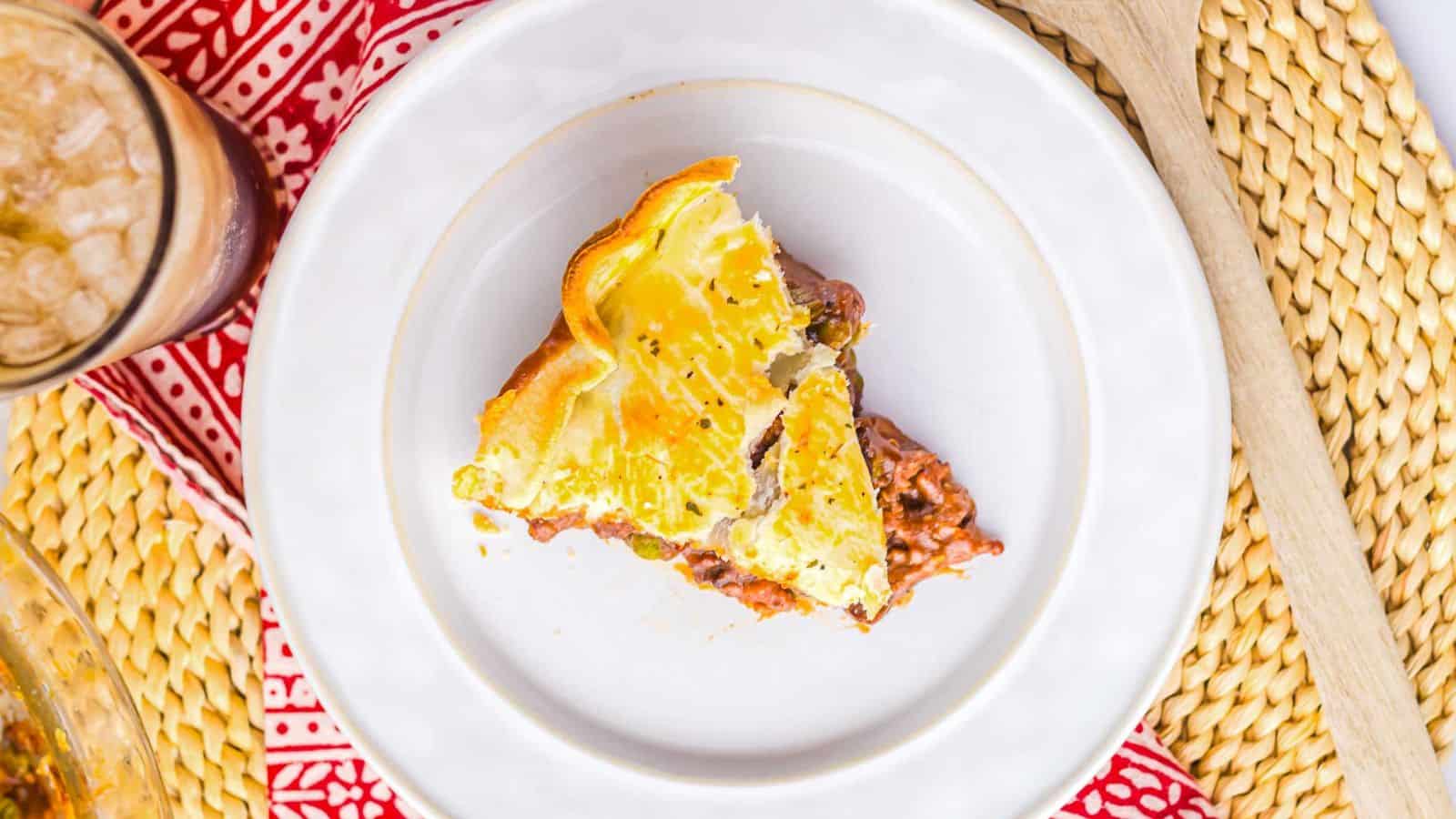 A close-up of a beef and vegetable pie with a golden, flaky crust. A triangular segment is removed, revealing chunks of beef and vegetables in a savory sauce inside the pie. The crust is lightly seasoned with herbs.