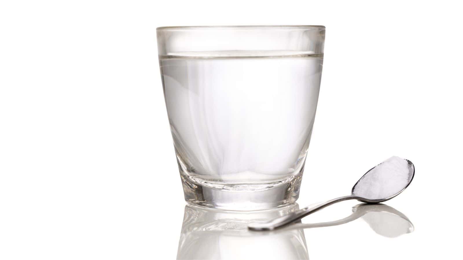 A clear glass filled with water is placed on a reflective surface. Beside it, a metal spoon holds a small mound of white powder, likely sugar or salt. The background is white, creating a clean and simple composition.