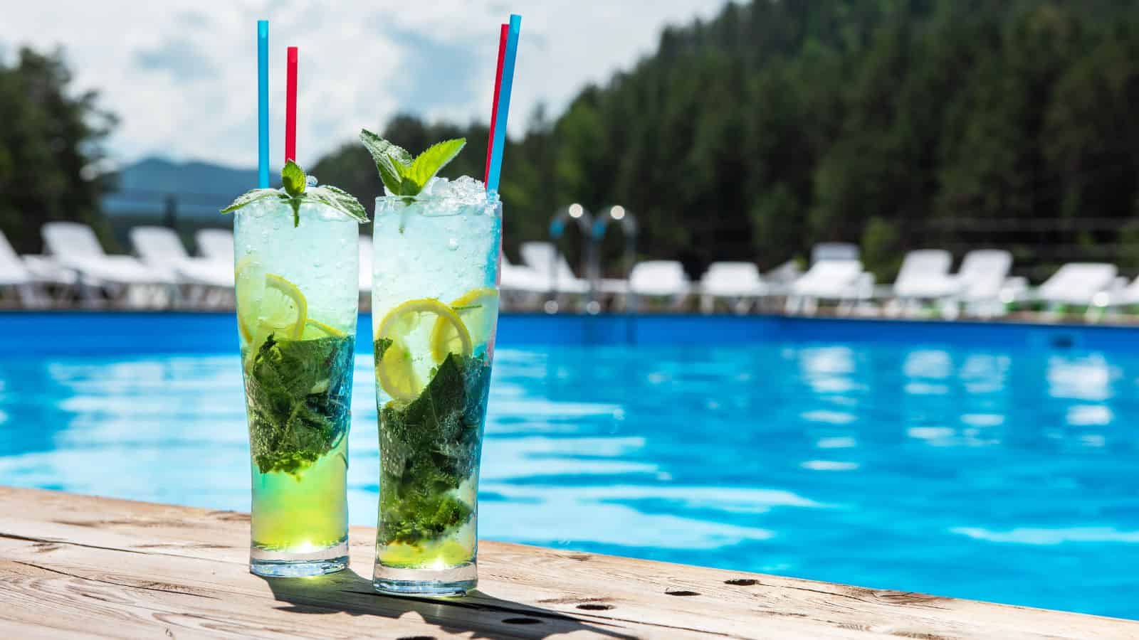 Two tall glasses filled with a mojito-style drink, containing mint leaves, lemon slices, and ice, are placed on a wooden poolside deck. Each glass has a red and blue straw. In the background, there is a clear blue pool and blurred greenery.