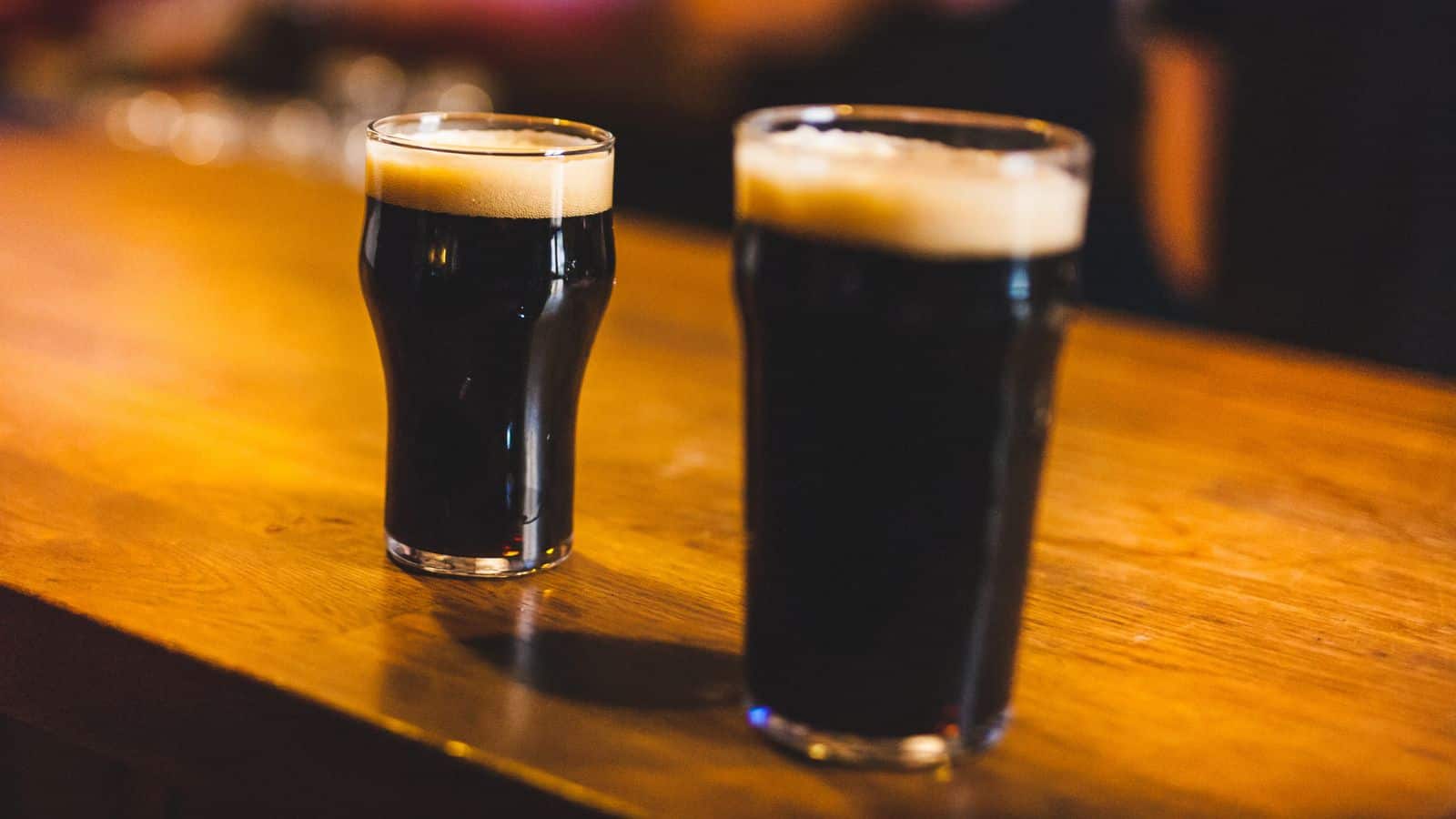 Two pint glasses filled with dark beer are placed on a wooden bar counter. The beer has a creamy foam head. The background is blurred, focusing on the drinks.