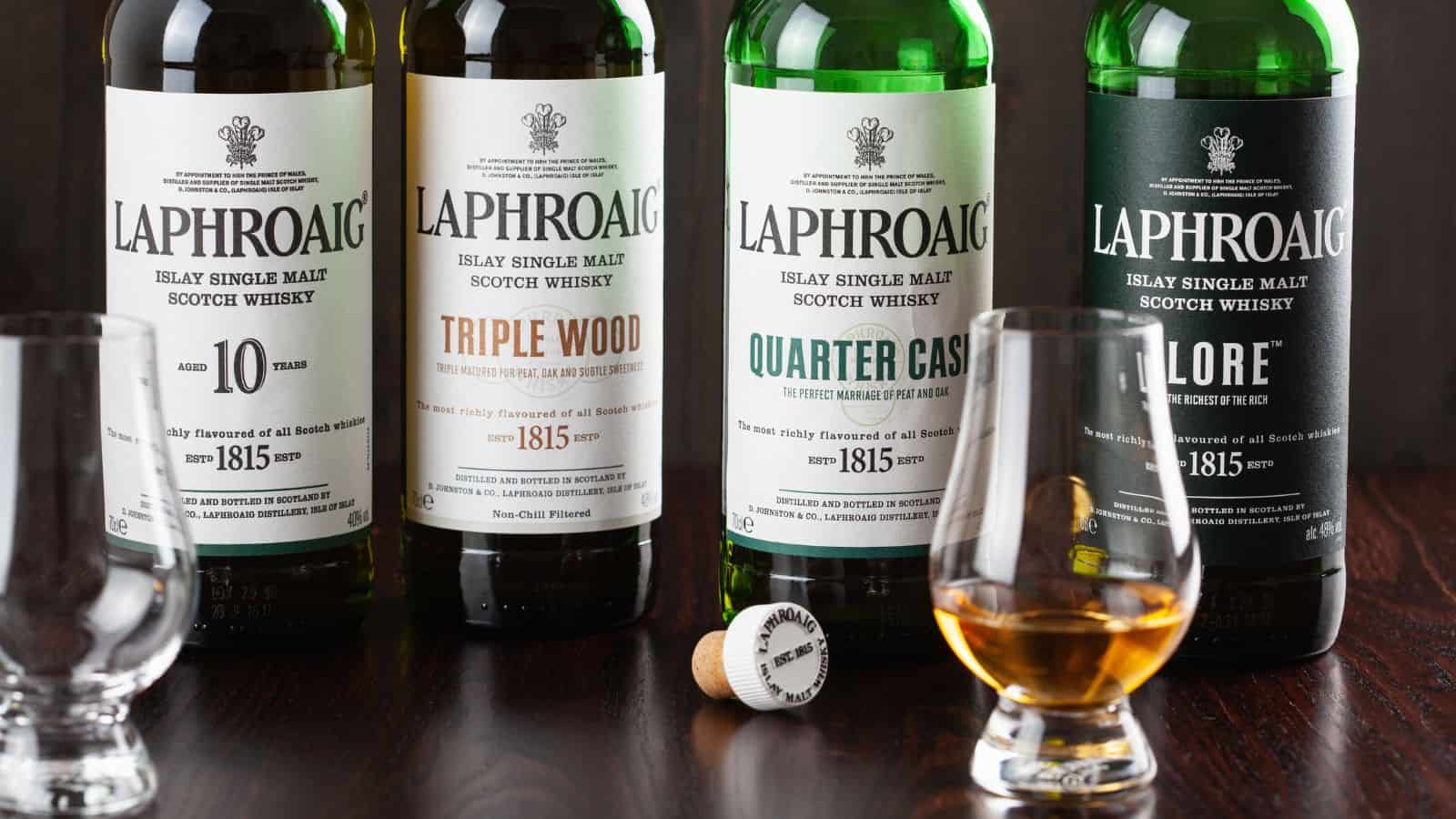 Four bottles of Laphroaig whisky are lined up, labeled as 10 Year Old, Triple Wood, Quarter Cask, and Lore. Two glasses are in front, one empty and one with whisky. A bottle cap and cork are placed nearby on a dark wooden surface.