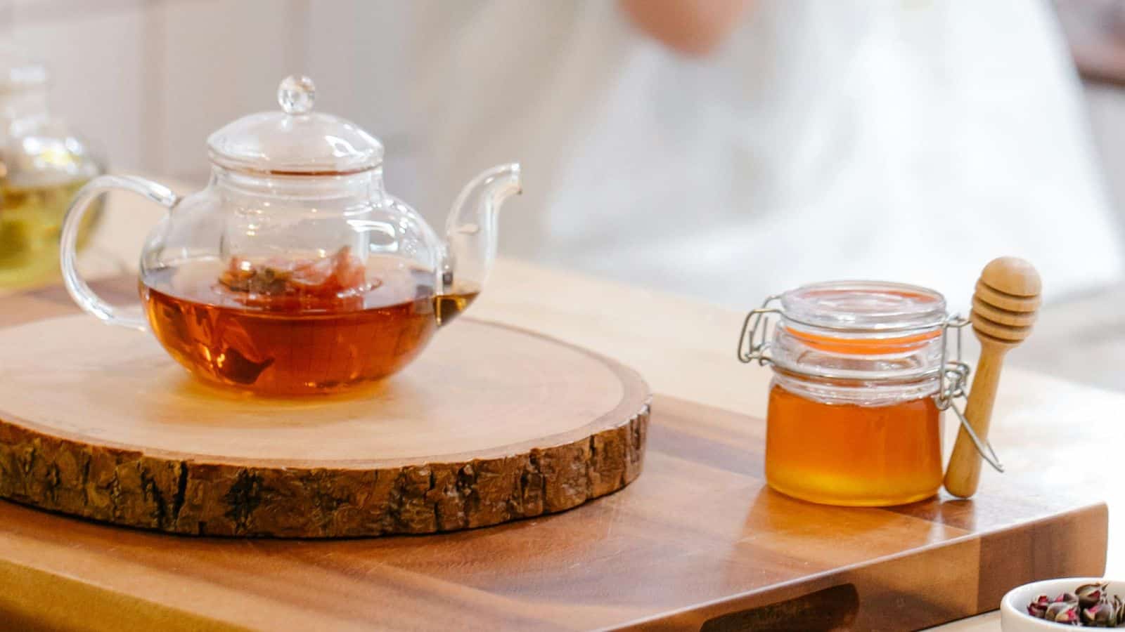 A glass teapot with tea sits on a wooden slice coaster. Next to it, a small jar of honey with a wooden dipper is placed on a wooden table. The background is softly blurred.