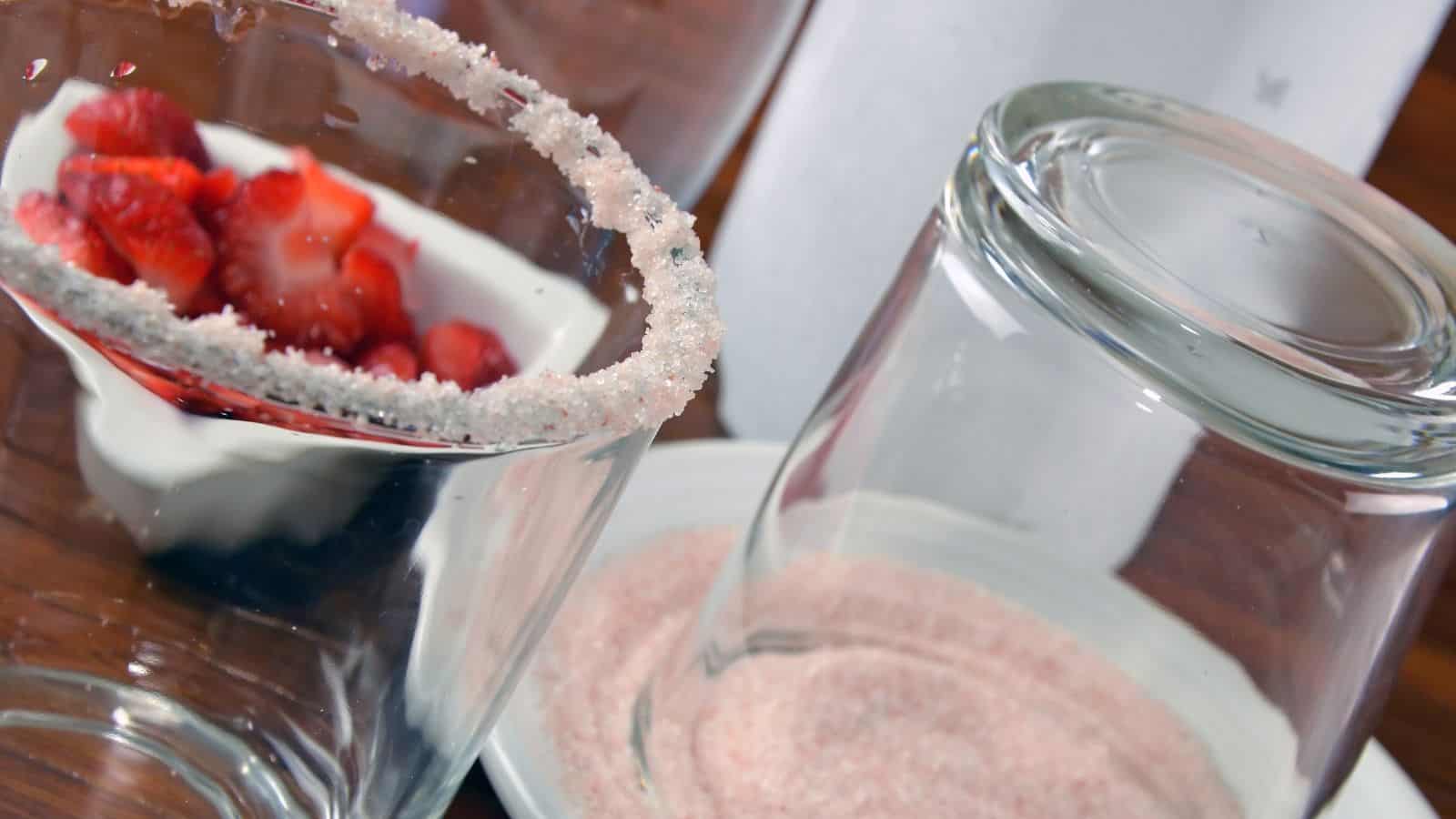 A glass is being rimmed with pink sugar, lying upside down on a plate containing the sugar. Nearby, a bowl holds sliced strawberries and cream, placed on a wooden surface.