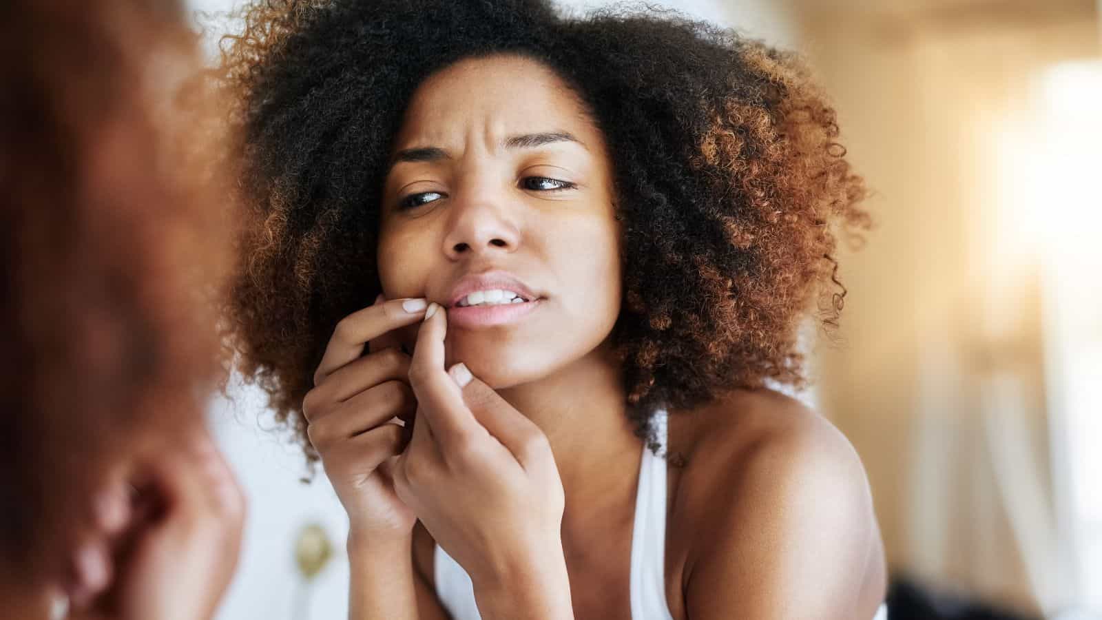 A person with curly hair is looking at their reflection in a mirror, examining their face and touching their chin. They appear to be checking for a blemish or similar skin condition. The lighting is soft and warm.