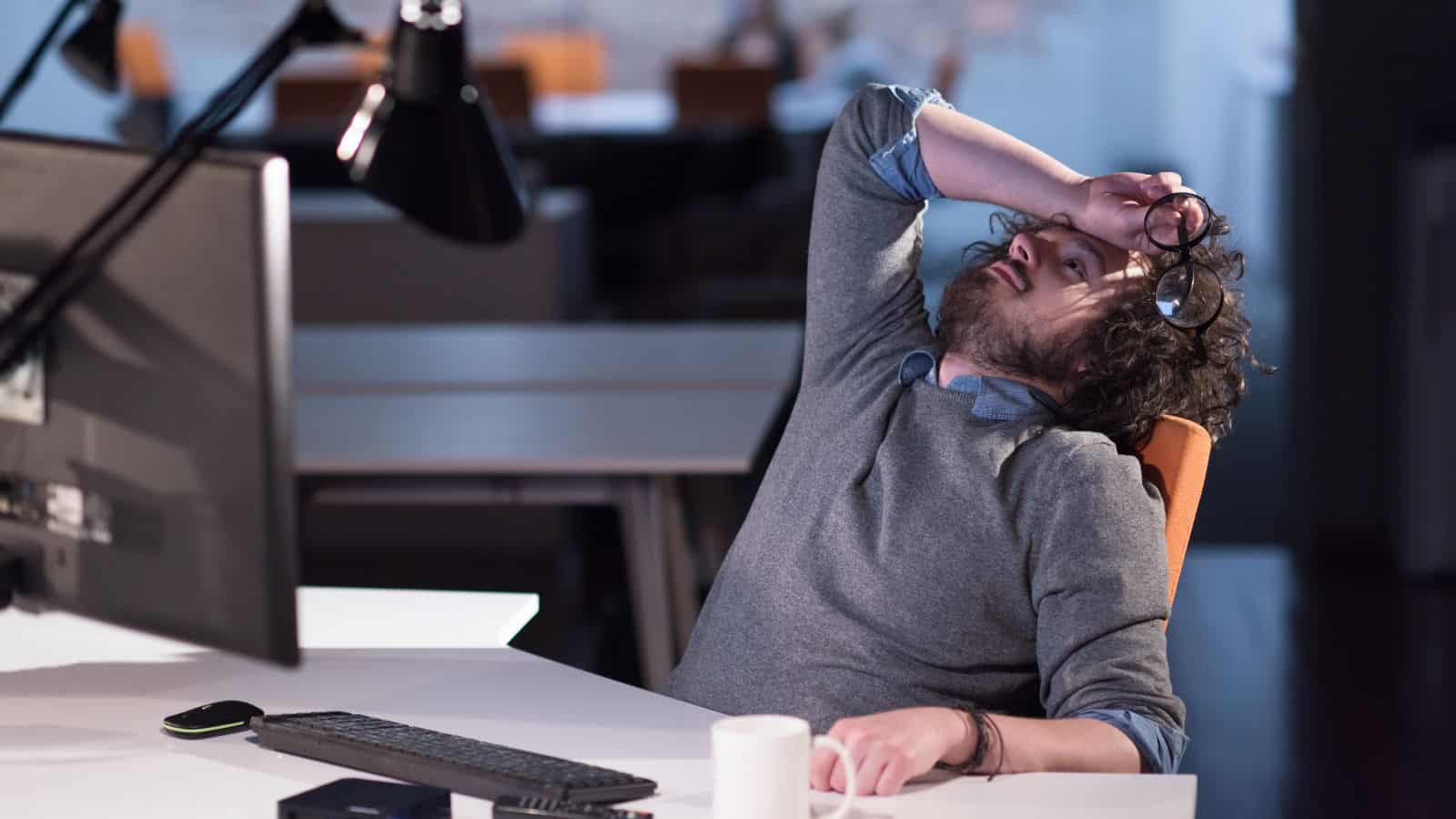 A person sits at a desk in an office, leaning back with their glasses held in one hand against their forehead. They appear tired or stressed. A computer monitor, a keyboard, a mug, and a closed notebook are on the desk.