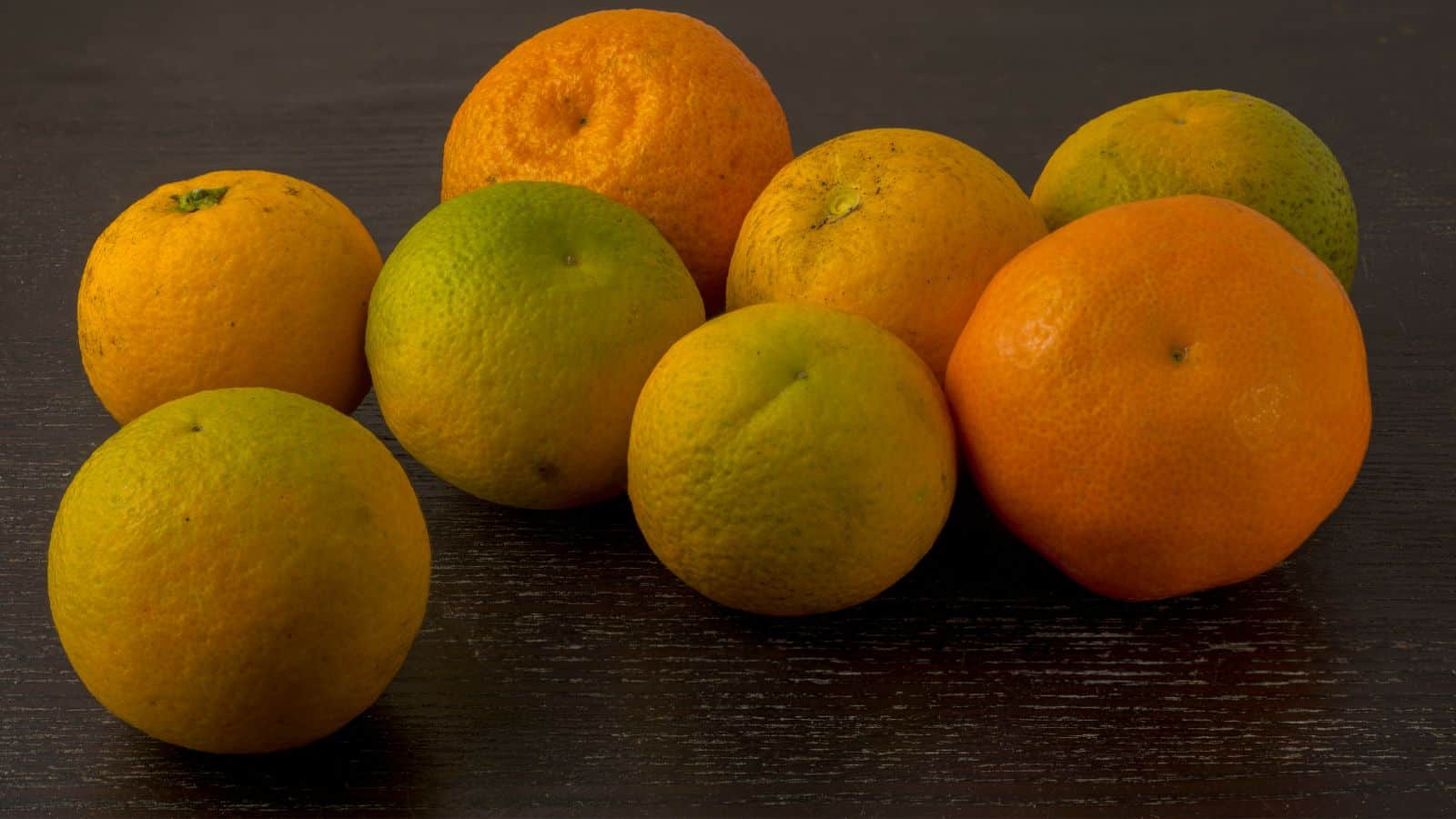 A group of eight citrus fruits, including oranges and possibly grapefruits, is arranged on a dark wooden surface. The fruits vary in shades of orange and green, and the surface shows some texture.