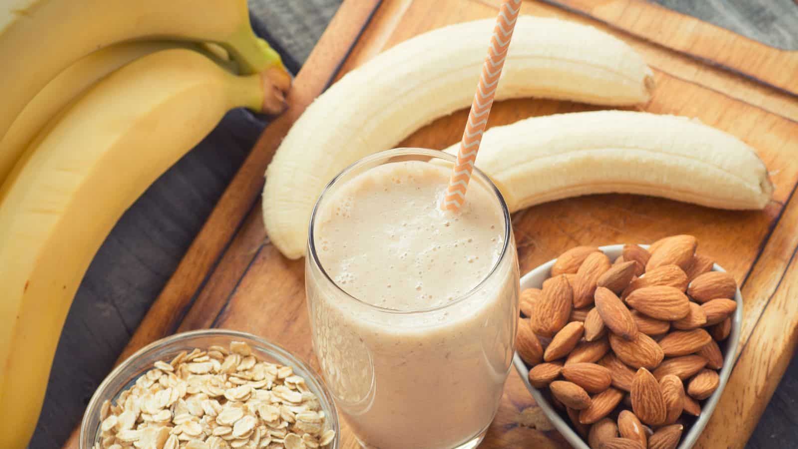 A glass of banana smoothie with a straw is on a wooden board. Beside it are whole and peeled bananas, a small bowl of almonds, and a bowl of oats. The setup suggests ingredients for making the smoothie.