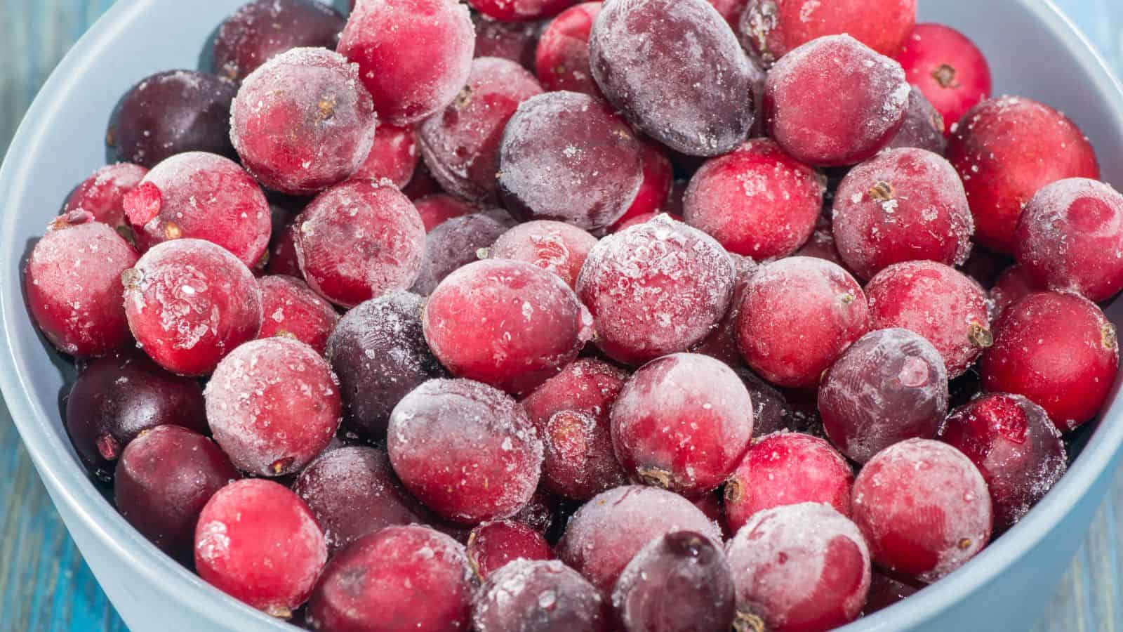 A bowl filled with frozen red cranberries, covered with a light layer of frost. The bowl is set against a blue wooden surface.
