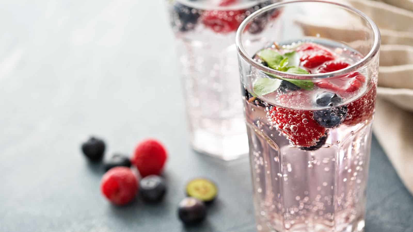 Two glasses of sparkling water filled with raspberries, blueberries, and mint leaves on a light gray surface. Berries are scattered nearby. The focus is on the front glass, and a napkin is partially visible to the right.