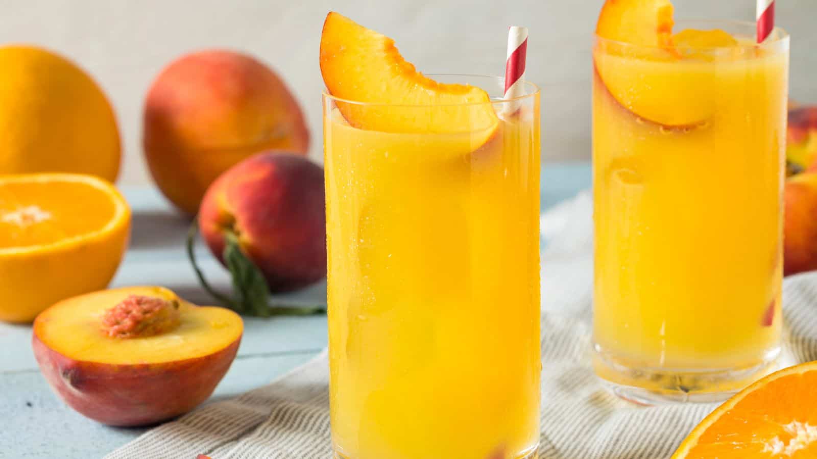 Two glasses of orange juice with peach slices and striped straws are on a white napkin. Surrounding them are whole peaches, an orange, and a halved peach on a blue surface.
