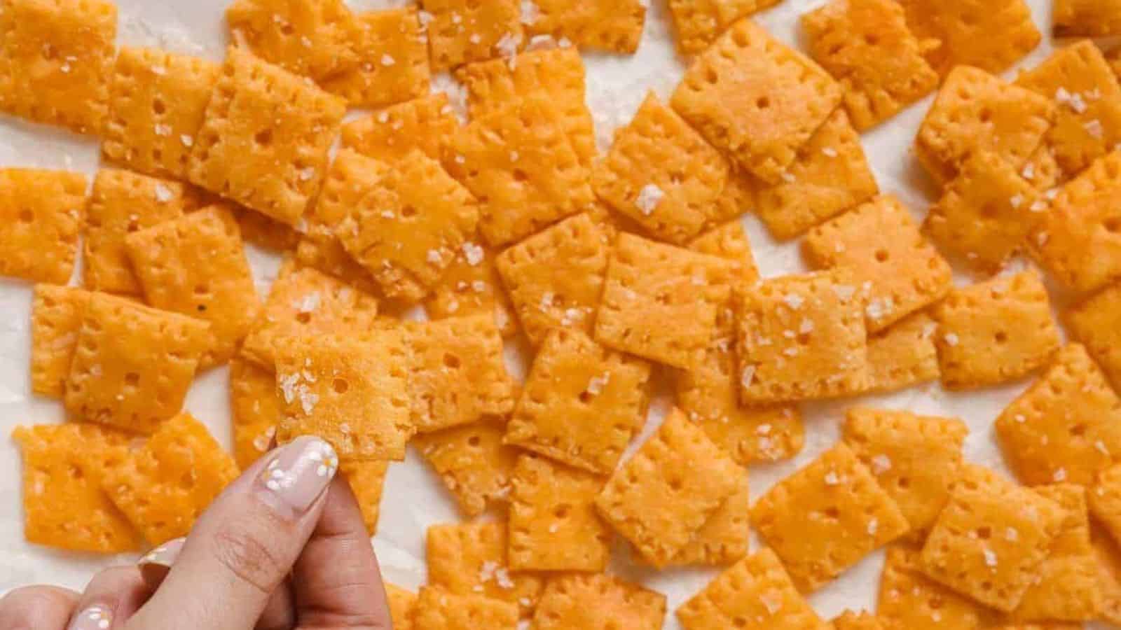 A hand holding a Cheez-It above a baking sheet of more crackers.