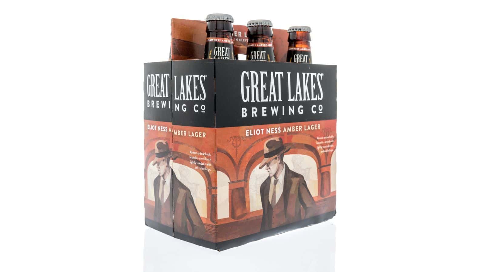 A six-pack of Great Lakes Brewing Co. Eliot Ness Amber Lager is displayed. The packaging features an illustration of a man in a trench coat and hat against an art deco backdrop. Four of the six bottles are visible with similar labels.