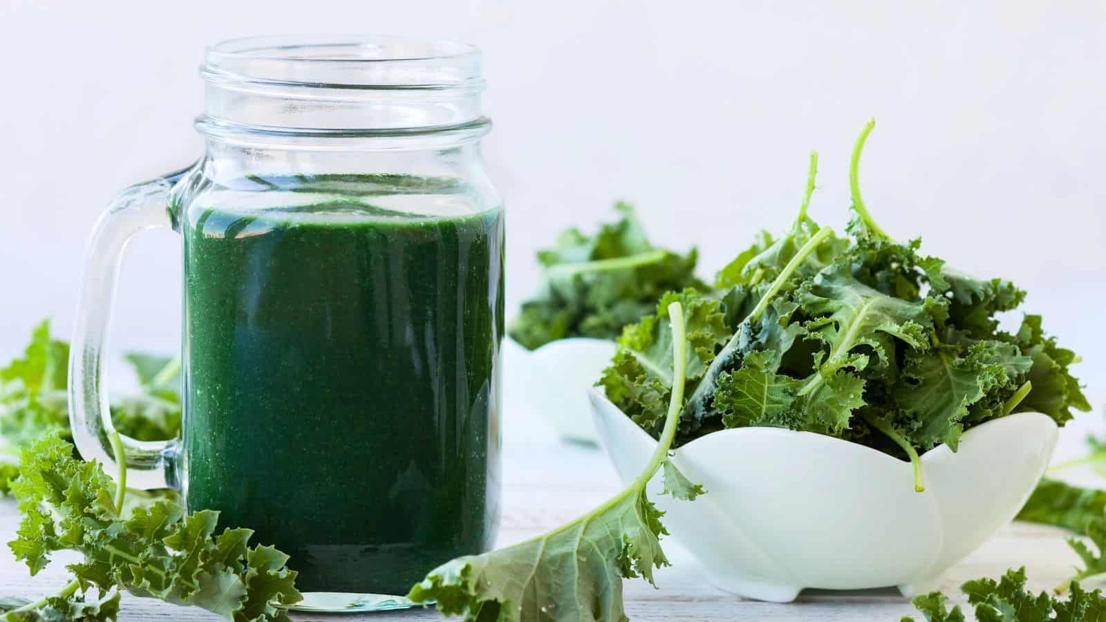 A clear glass mug filled with dark green smoothie sits on a light surface. Next to it, a white bowl holds fresh kale leaves. Additional kale leaves are scattered around the mug and bowl. The background is softly blurred.
