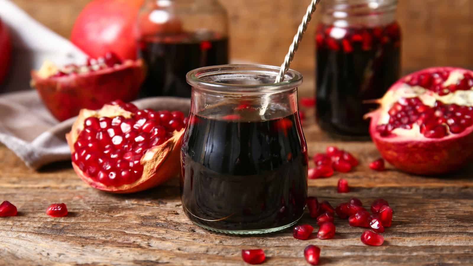 A glass jar filled with dark juice sits on a wooden surface with a stainless steel straw inside. Surrounding it are open pomegranates, revealing bright red seeds, and additional jars filled with juice. Loose seeds are scattered on the surface.