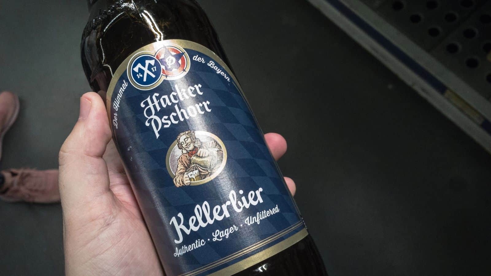 A person holds a bottle of Hacker Pschorr Kellerbier. The label is dark blue with a diamond pattern and features an illustration of a man. Text on the label includes "authentic," "lager," and "unfiltered." Shoes are visible in the background.