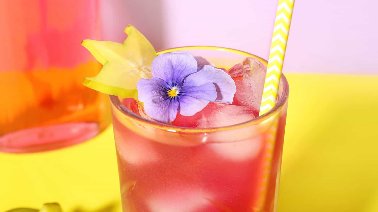 A refreshing pink drink in a glass with ice cubes, garnished with a purple flower and a slice of starfruit. The drink has a yellow and white striped straw. The background features a pink and yellow gradient surface.