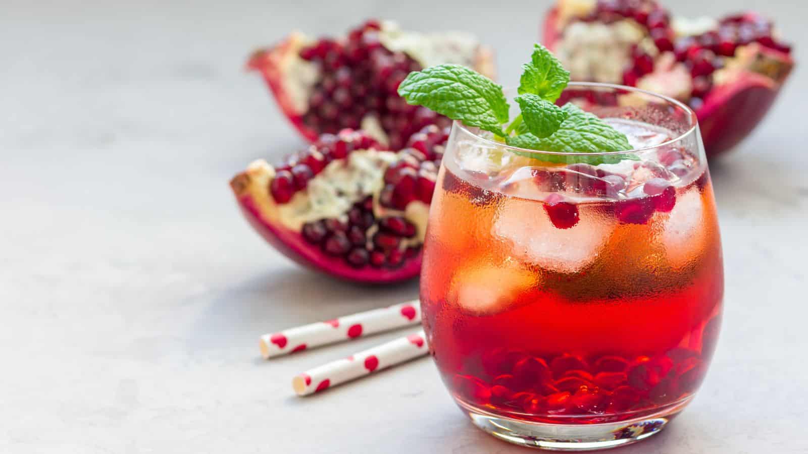 A glass of red pomegranate juice with ice cubes is garnished with a sprig of mint. In the background, two half pomegranates with seeds visible are placed on a light gray surface. Two red and white straws lie next to the glass.
