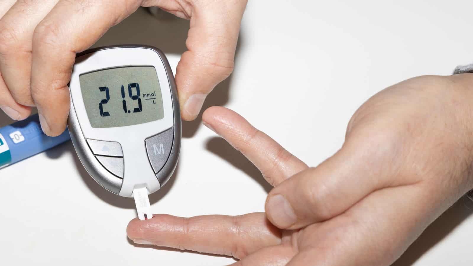 A hand is holding a glucose meter showing a reading of 21.9 mmol/L. Another hand is positioned with a finger near the test strip, indicating a blood sugar testing process. The background is a plain white surface.