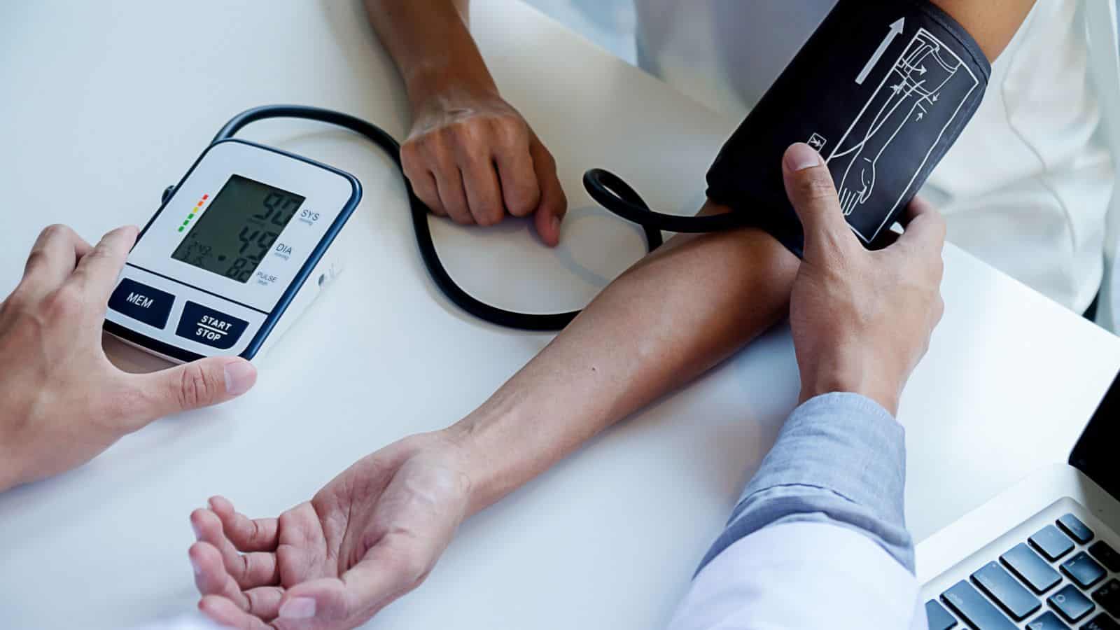 A person is having their blood pressure taken using a digital monitor. The cuff is wrapped around their left arm. Another person is operating the device. A laptop is partially visible on the table.