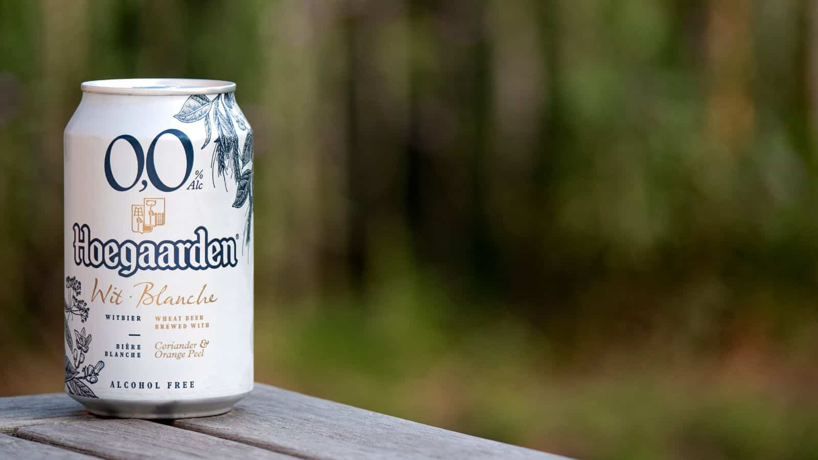 A can of Hoegaarden 0.0% Alcohol-Free Wit Blanche sits on a wooden surface with a blurred green background. The white can is decorated with blue floral graphics and text indicating it is a wheat beer brewed with coriander and orange peel.