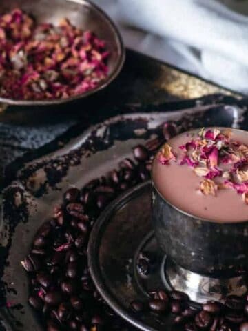 A cup filled with pinkish liquid topped with dried rose petals sits on a dark tray surrounded by coffee beans. Nearby, a small dish contains cardamom pods, and another bowl holds more dried rose petals. A white cloth is partially visible.