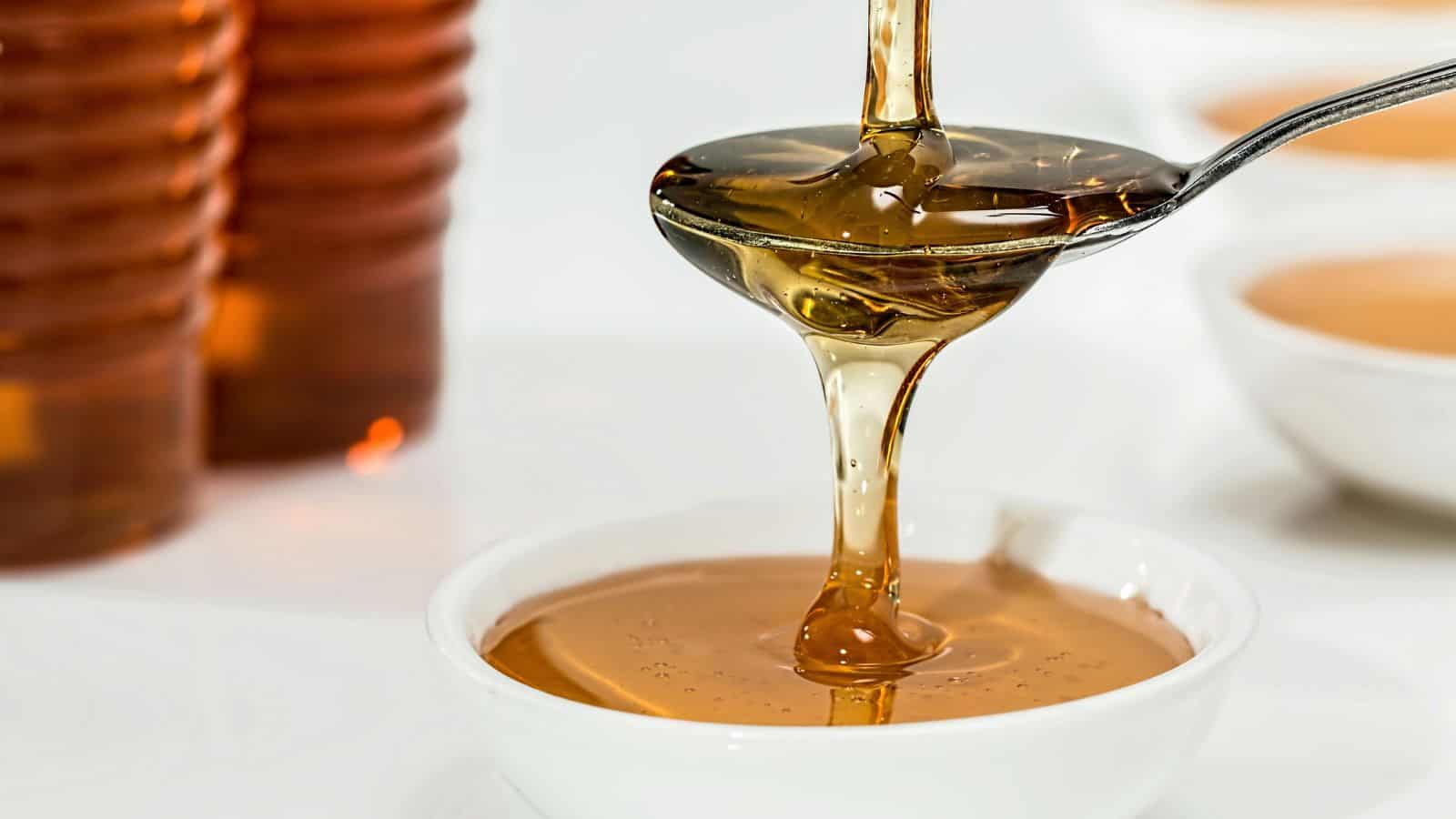 A spoon is shown pouring golden honey into a white bowl. In the background, there are blurred honey containers. The honey is thick and glossy, cascading in a smooth stream from the spoon.