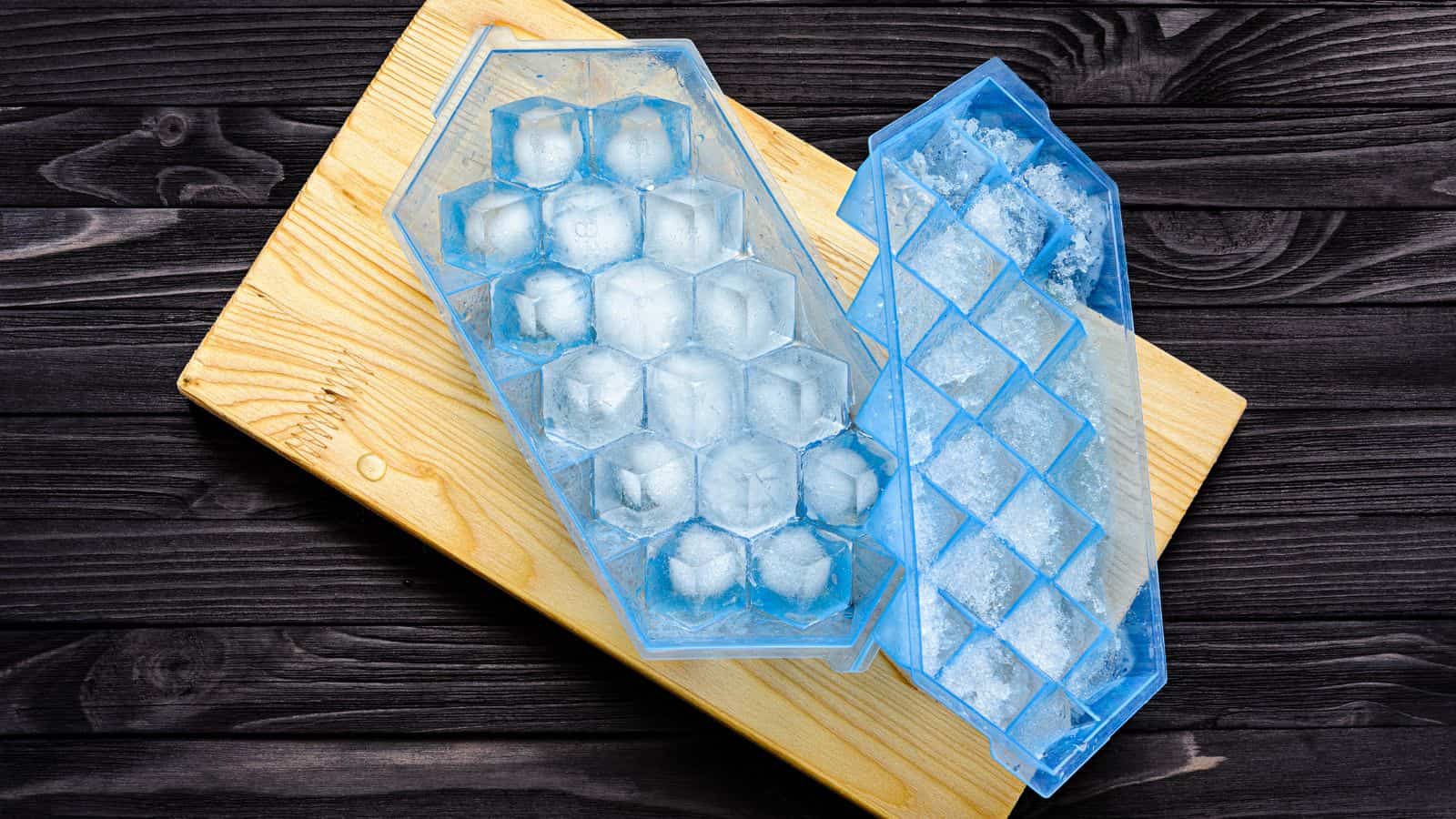 Two blue silicone ice cube trays, one filled with ice cubes and the other partially empty, are placed on a light wooden cutting board set against a dark wooden background.