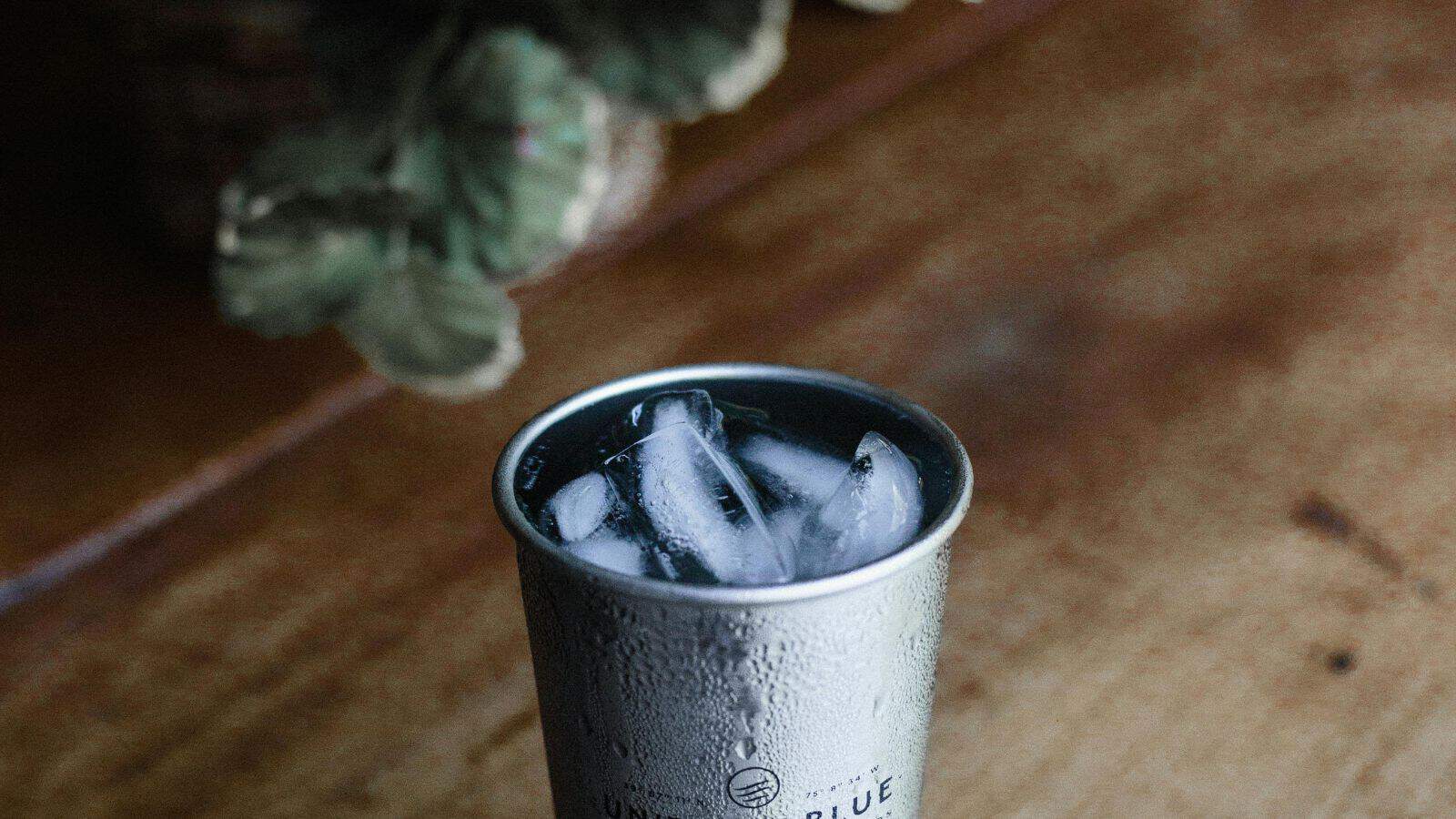 A metal cup with droplets on its surface is filled with ice cubes. The cup is placed on a wooden table, and there are blurred green leaves in the background.
