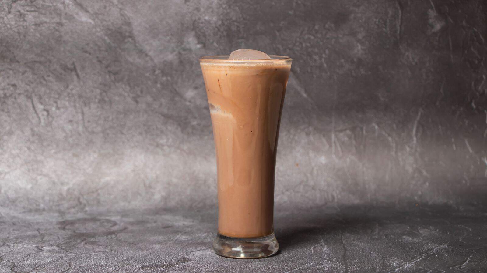 A tall glass filled with iced coffee sits on a textured gray surface. The drink is a light brown color and contains visible ice cubes. The background is a gray, textured wall.