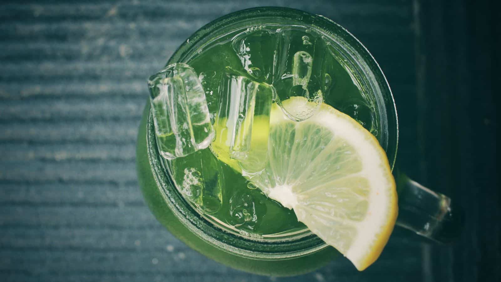 A top view of a glass mug filled with green liquid and ice cubes. A lemon slice is placed on the rim of the mug. The background is a textured, dark surface.