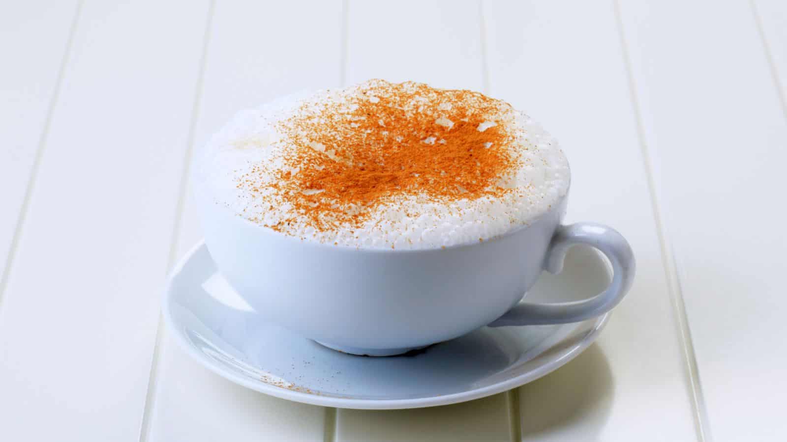 A cup of cappuccino topped with a generous amount of cinnamon sits on a white saucer. The drink is placed on a white table, and the cinnamon forms a light brown layer over the frothy milk.