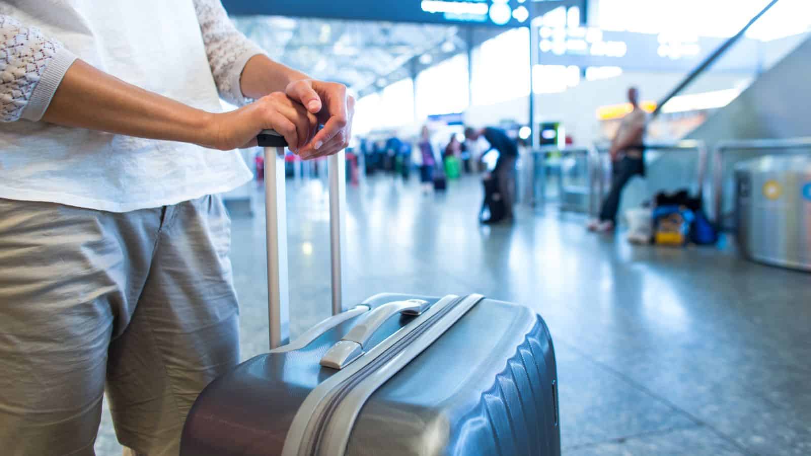 Person standing at an airport terminal with hands resting on the handle of a suitcase. The background is blurred, showing other travelers and departure signage. The atmosphere is busy with visible motion and activity.