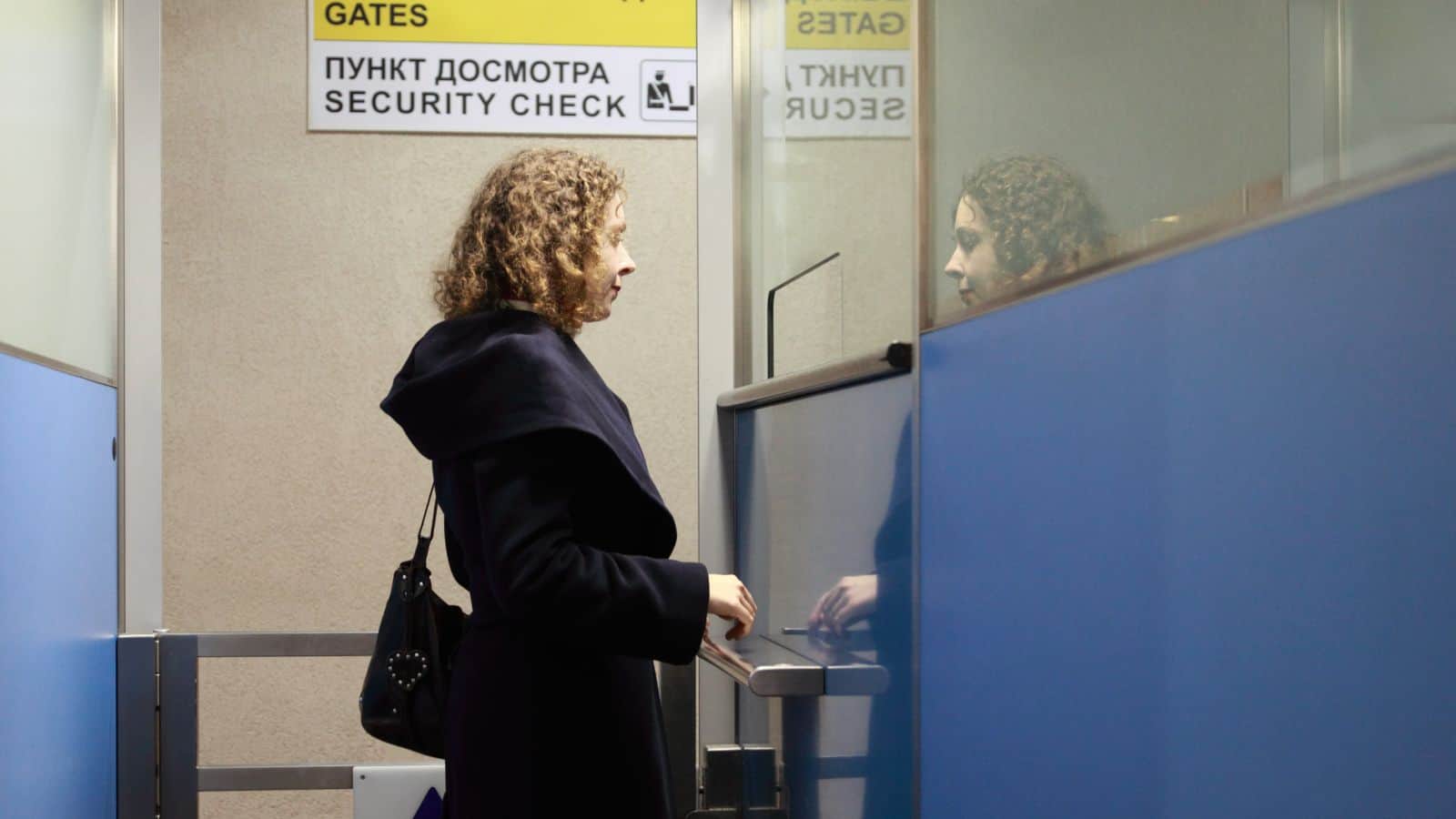 A person with curly hair and a dark coat stands at a security check entrance, holding an access card. A bilingual sign above reads "Gates" and "Security Check" in English and Russian. Blue panels line the hallway.